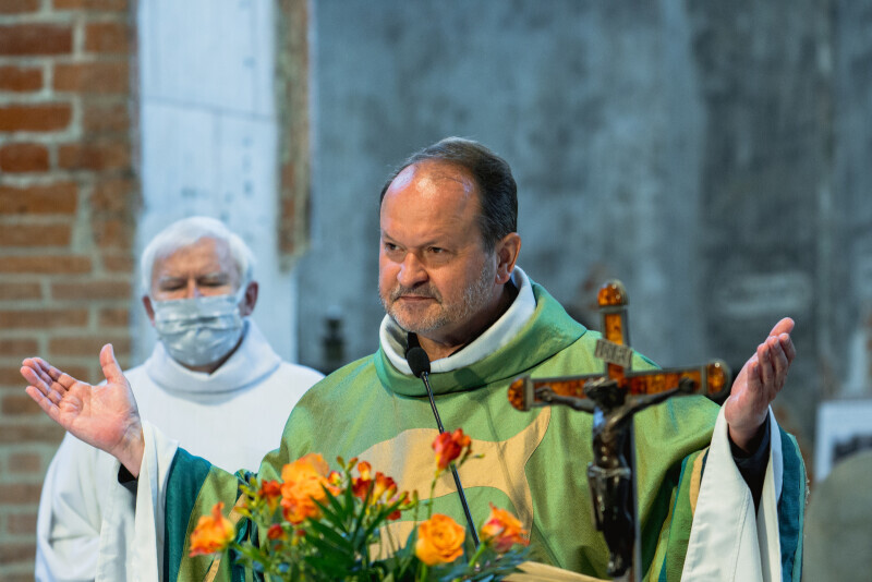 Ksiądz Krzysztof Niedałtowski ubrany w zieloną szatę, podczas odprawnia mszy święcej. Na pierwszym planie pomarańczowe kwiaty i krzyż stojące na ołtarzu. Na drygim planie zakonnik ubrany w białą szatę i maseczkę.